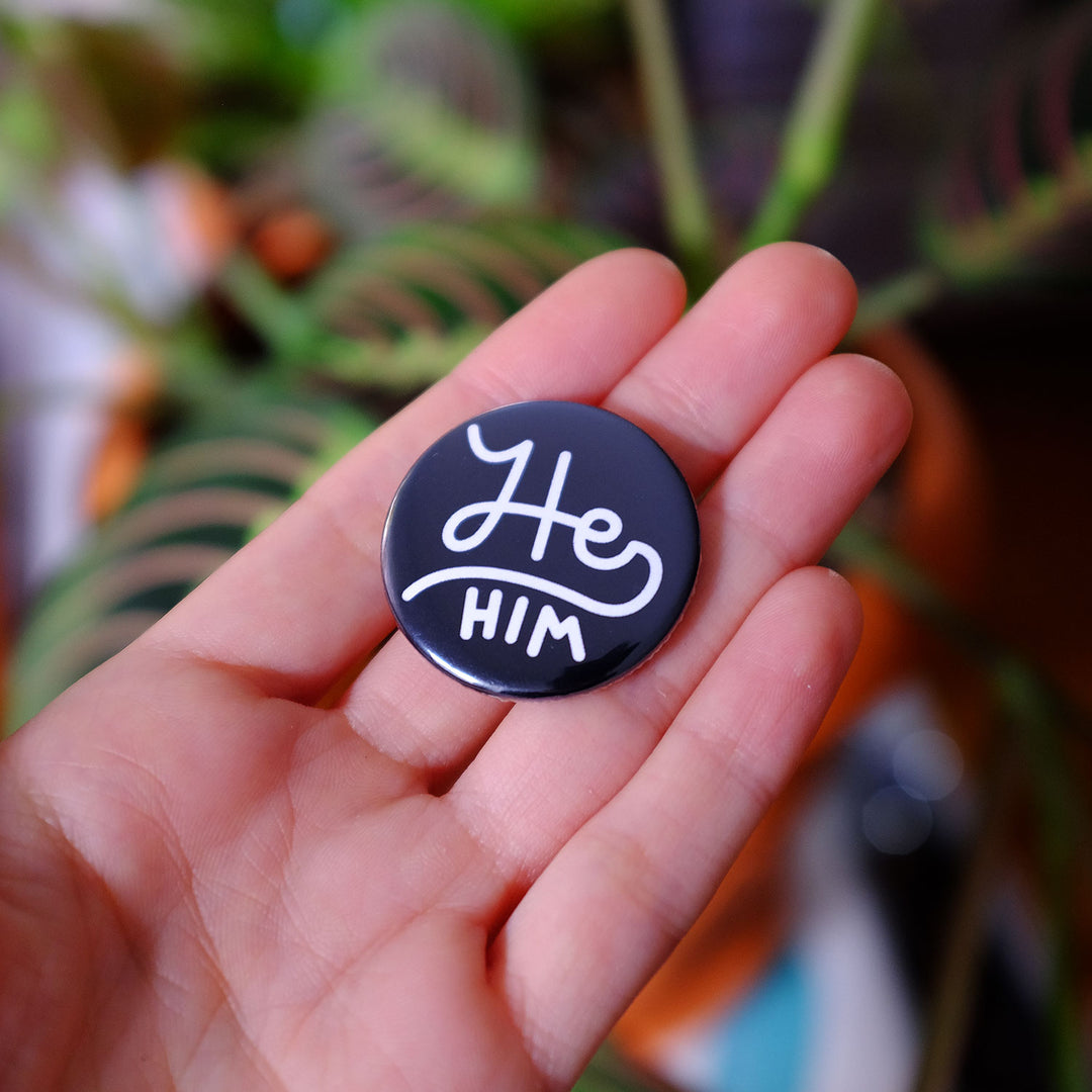 Hand holding the He/him Pronouns Button by Bianca Designs.