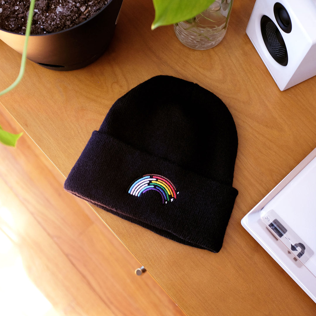 Inclusive Rainbow Pride Beanie in Black by Bianca Designs. The beanie is laying flat on a wooden table.