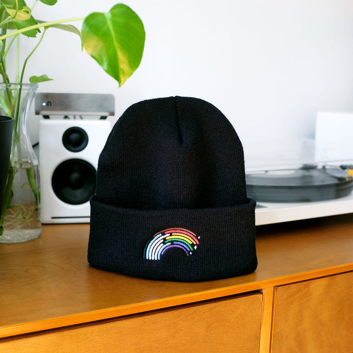 Inclusive Rainbow Pride Beanie in Black by Bianca Designs. The beanie is propped up on a wooden table.