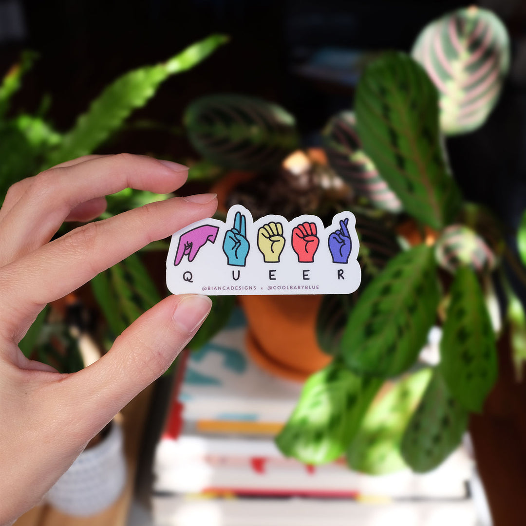 Hand holding the Queer ASL Fingerspell Pride Sticker by Bianca Designs & Gregor Lopes. Plants are in the background.