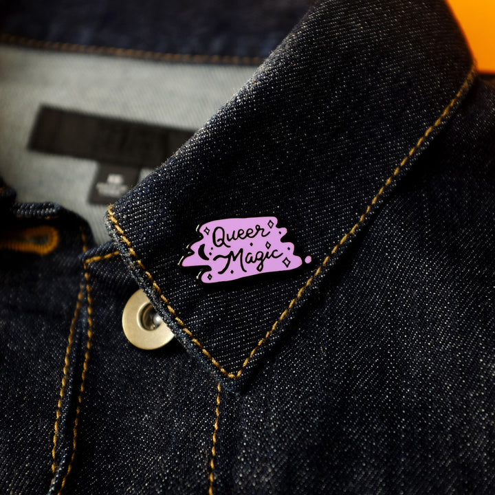 Queer Magic Pin, in Purple and Black, by Bianca Designs. The pin is pinned to a denim jacket's collar.