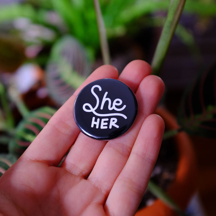 Hand holding the She/her Pronouns Button by Bianca Designs.